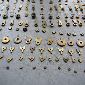 May include: A collection of small, vintage brass watch parts, including gears, levers, and various shaped components. The parts are arranged on a gray surface, showcasing their intricate designs and aged patina. The components vary in size and shape.