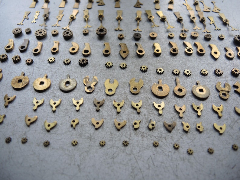 May include: A collection of small, vintage brass watch parts, including gears, levers, and various shaped components. The parts are arranged on a gray surface, showcasing their intricate designs and aged patina. The components vary in size and shape.