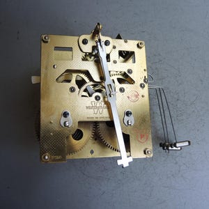 May include: A brass clock movement mechanism featuring exposed gears, a central vertical pendulum rod, and a series of thin metal chime hammers. The plate is stamped with the text WESTERSTRAND and MADE IN SWEDEN, along with a circular red logo.