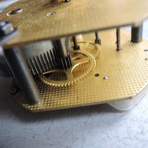 Op de afbeelding: Close-up van een uurwerk met messing tandwielen en platen. De tandwielen zijn in elkaar grijpend en de platen hebben een gestructureerd oppervlak. Het uurwerk is waarschijnlijk vintage of antiek en wordt gerepareerd.