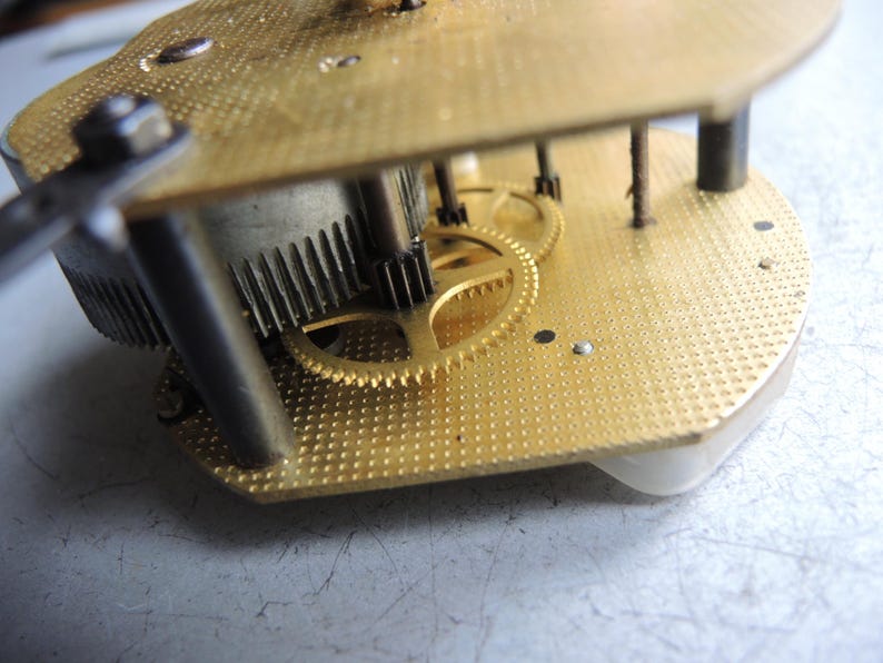 Op de afbeelding: Close-up van een uurwerk met messing tandwielen en platen. De tandwielen zijn in elkaar grijpend en de platen hebben een gestructureerd oppervlak. Het uurwerk is waarschijnlijk vintage of antiek en wordt gerepareerd.
