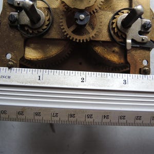 May include: Close-up of a clock mechanism with brass gears and metal components. A metal ruler, marked in inches, is placed across the mechanism. The ruler shows measurements up to 4 inches.