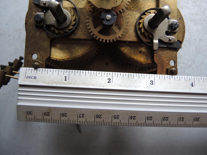 May include: Close-up of a clock mechanism with brass gears and metal components. A metal ruler, marked in inches, is placed across the mechanism. The ruler shows measurements up to 4 inches.