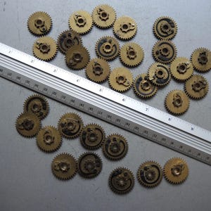 May include: A collection of small, brass-colored gears with intricate details, scattered on a gray surface. A metal ruler with inch markings is placed diagonally across the gears, providing scale. The gears appear to be vintage or antique.