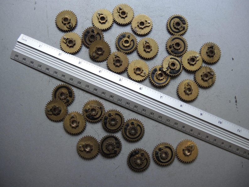 May include: A collection of small, brass-colored gears with intricate details, scattered on a gray surface. A metal ruler with inch markings is placed diagonally across the gears, providing scale. The gears appear to be vintage or antique.