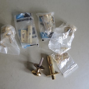 May include: An assortment of new, brass-colored door hardware components, including doorknobs, hinges, and latches, are packaged in clear plastic bags. The hardware is arranged on a light gray surface, ready for installation.