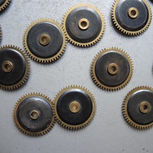 May include: A collection of vintage watch gears. Each gear features a brass-colored toothed outer ring and a dark, circular center with a central hole. The gears are scattered across a light gray surface.