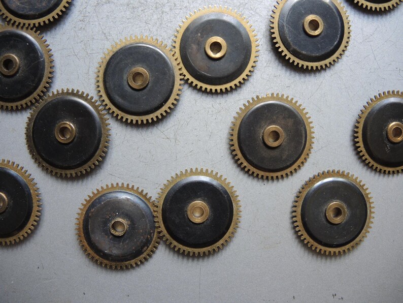 May include: A collection of vintage watch gears. Each gear features a brass-colored toothed outer ring and a dark, circular center with a central hole. The gears are scattered across a light gray surface.