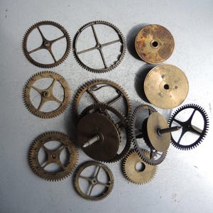 May include: A collection of various antique clock gears and components, in shades of gold and bronze. The gears vary in size and design, with circular and spoked patterns. Some gears have a dark, aged patina.