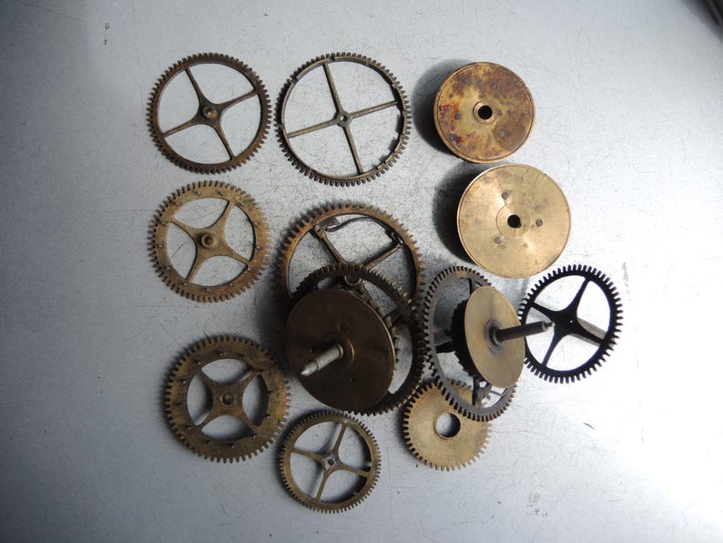 May include: A collection of various antique clock gears and components, in shades of gold and bronze. The gears vary in size and design, with circular and spoked patterns. Some gears have a dark, aged patina.