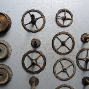 May include: An assortment of vintage clock gears and components in various sizes and shapes. The gears are made of aged metal, displaying a range of sizes and designs, including toothed wheels and circular plates. The arrangement suggests a collection of antique clock parts.