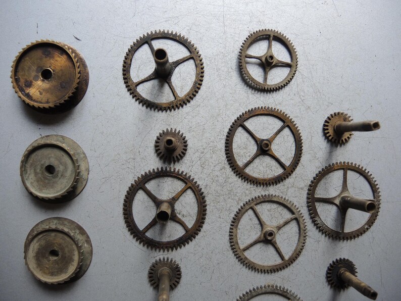 May include: An assortment of vintage clock gears and components in various sizes and shapes. The gears are made of aged metal, displaying a range of sizes and designs, including toothed wheels and circular plates. The arrangement suggests a collection of antique clock parts.