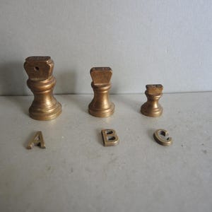 May include: Three antique brass chess pieces and three small brass letters, A, B, and C, are arranged on a light grey surface. The chess pieces are in the shape of a rook, with a square top and a cylindrical base.