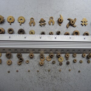 May include: A collection of small, brass-colored gears and mechanical components of various shapes and sizes. A metal ruler with inch markings is placed across the top of the gears. The gears range from approximately 0.1 to 0.75 inches in diameter.