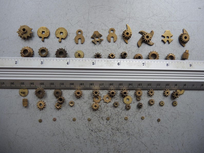 May include: A collection of small, brass-colored gears and mechanical components of various shapes and sizes. A metal ruler with inch markings is placed across the top of the gears. The gears range from approximately 0.1 to 0.75 inches in diameter.
