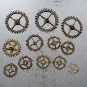 May include: A selection of bronze-coloured gears of different sizes, each with toothed edges and central hubs. The gears are arranged on a light grey surface, highlighting their intricate designs and mechanical details. The gears vary in size, with some having four spokes and others five.