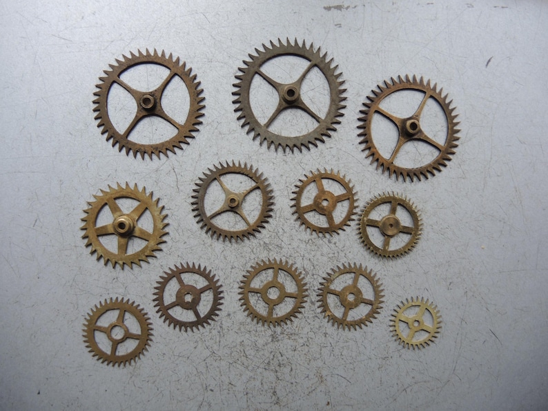 May include: A selection of bronze-coloured gears of different sizes, each with toothed edges and central hubs. The gears are arranged on a light grey surface, highlighting their intricate designs and mechanical details. The gears vary in size, with some having four spokes and others five.