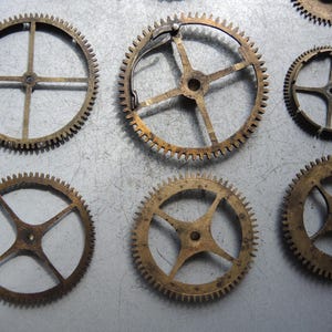 May include: A collection of antique brass gears of varying sizes. The gears have a circular shape with toothed edges and internal spokes. The metal has a tarnished, aged appearance, suggesting vintage or reclaimed components.