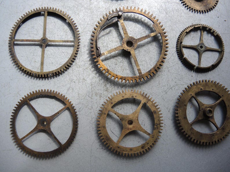 May include: A collection of antique brass gears of varying sizes. The gears have a circular shape with toothed edges and internal spokes. The metal has a tarnished, aged appearance, suggesting vintage or reclaimed components.