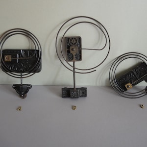 Puede incluir: Tres piezas antiguas de reloj se exhiben sobre un fondo neutro. Las piezas son de metal oscuro y presentan diseños intrincados. Una es circular, otra rectangular y la tercera cuadrada con una espiral.