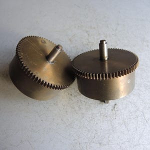 May include: Two brass gears with toothed edges and central shafts. The gears are cylindrical with a circular top and bottom. The gears are a warm, metallic colour and appear to be vintage or antique.