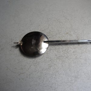 Op de afbeelding: Een glanzende, zilverkleurige pendel met een platte, rechthoekige verlenging. De ronde bob is bevestigd aan een dunne staaf, en een klein draadje is zichtbaar aan de bovenkant. Het object is waarschijnlijk een uurwerkonderdeel.
