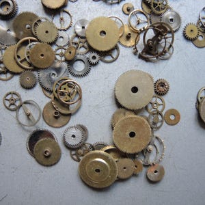 May include: A collection of various sized watch gears in brass and silver tones. The gears are scattered across a light gray surface, showcasing their intricate designs and mechanical details. Some gears have a central hole.