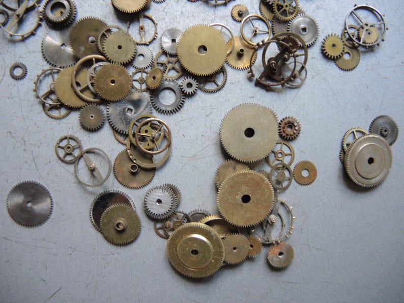 May include: A collection of various sized watch gears in brass and silver tones. The gears are scattered across a light gray surface, showcasing their intricate designs and mechanical details. Some gears have a central hole.