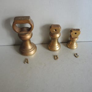 May include: Three antique brass weights of different sizes. The largest has a handle, the others have a flat top with a hole. Each weight is labelled with a letter: A, B, and C. The weights are displayed on a light grey surface.