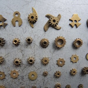 May include: A collection of small, brass-colored gears and mechanical components. Various shapes and sizes are visible, including circular, star-shaped, and uniquely designed pieces, scattered on a gray surface. These parts appear to be from a clock or watch.
