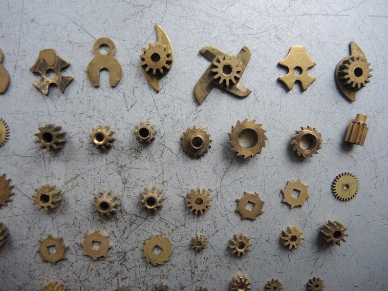 May include: A collection of small, brass-colored gears and mechanical components. Various shapes and sizes are visible, including circular, star-shaped, and uniquely designed pieces, scattered on a gray surface. These parts appear to be from a clock or watch.