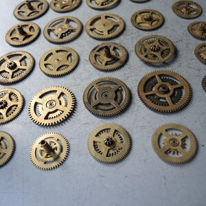 May include: A collection of small, brass-colored gears of varying sizes. The gears have intricate teeth and central hubs, suggesting they are components of a mechanical device. The arrangement is scattered, with some gears overlapping.