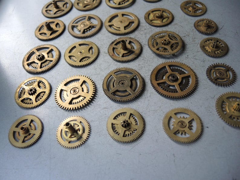 May include: A collection of small, brass-colored gears of varying sizes. The gears have intricate teeth and central hubs, suggesting they are components of a mechanical device. The arrangement is scattered, with some gears overlapping.