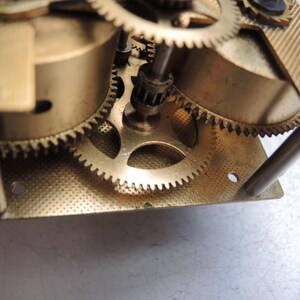 May include: Close-up of a clock mechanism with multiple brass gears and components. The gears are meshed together, and the metal has a gold tone. The image shows the intricate details of the clock's internal workings.