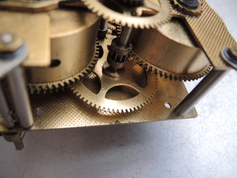May include: Close-up of a clock mechanism with multiple brass gears and components. The gears are meshed together, and the metal has a gold tone. The image shows the intricate details of the clock's internal workings.