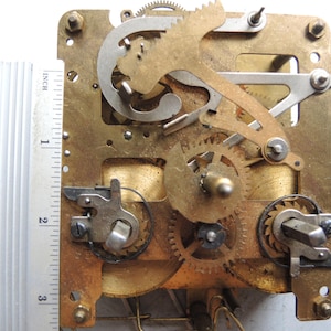 May include: Close-up of a clock mechanism with brass gears, levers, and a metal frame. A ruler shows the mechanism is approximately 3 inches (7.6 cm) wide. The gears are interlocked, and the frame has mounting holes.