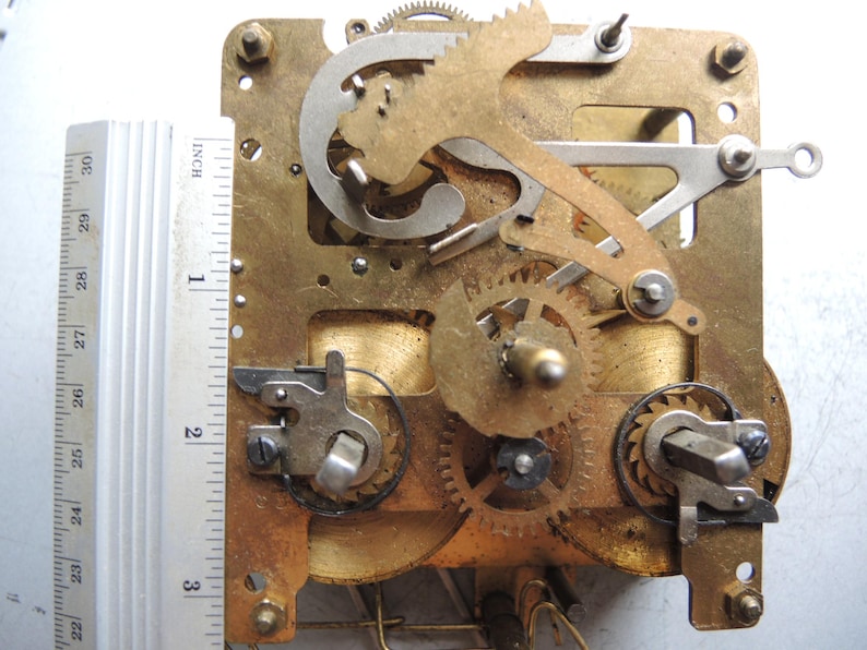May include: Close-up of a clock mechanism with brass gears, levers, and a metal frame. A ruler shows the mechanism is approximately 3 inches (7.6 cm) wide. The gears are interlocked, and the frame has mounting holes.