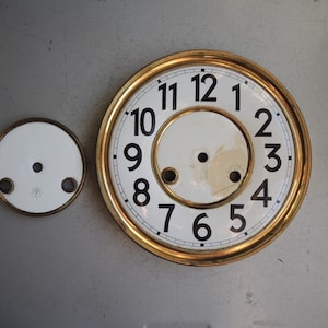 May include: Two antique clock faces. The larger face has a gold-colored rim, a white background, and black numerals. The smaller face is white with a gold-colored rim. Both have holes for clock hands.