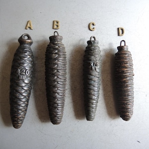 May include: Four antique metal clock weights, shaped like pine cones, with varying sizes and surface textures. Each weight has a loop at the top for hanging. One weight has the number "420" stamped on it. The weights are labelled A, B, C, and D.