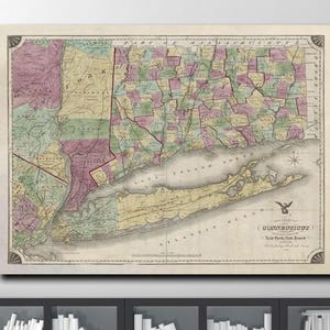 May include: A framed vintage map of Connecticut and Long Island, featuring a detailed depiction of counties in various colors. The map is displayed on a wall above a modern white chair and black bookshelf.