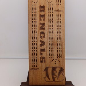 May include: Wooden cribbage board with the word "BENGALS" vertically engraved. The board features scoring holes, a team logo, and the words "START", "FINISH", and "SCORE". The board is displayed on a small wooden stand.