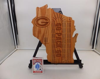 May include: Wooden cribbage board in the shape of Wisconsin, featuring the Green Bay Packers logo and the text "GO PACK GO." A deck of Bicycle playing cards is in front of the board. The board is displayed on a black stand.
