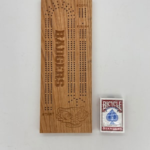 May include: A wooden cribbage board with the word "BADGERS" engraved vertically. The board has scoring holes and a Badger logo. A standard Bicycle playing card deck is next to the board.