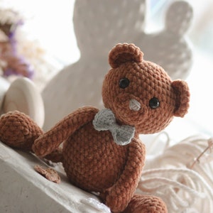 Könnte beinhalten: Ein brauner gehäkelter Teddybär mit einer hellblauen Schleife. Der Bär sitzt auf einer weißen Oberfläche mit einem weißen Seil und einem getrockneten Blumenstängel im Hintergrund.