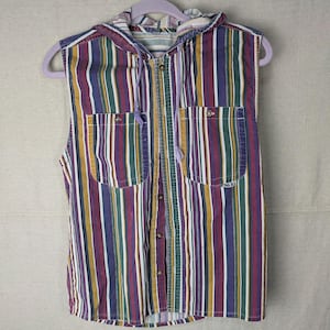 May include: Sleeveless hooded shirt with vertical stripes in shades of purple, green, yellow, and white. The shirt has two front pockets, a button-up closure, and a drawstring hood. The shirt is hanging on a lavender hanger.