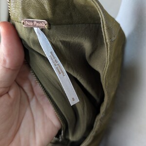 May include: Close-up of an olive green garment with a zipper and a tag that reads "Free People". The tag also includes the text "FABRIQUE EN CHINE". The garment is size 8.