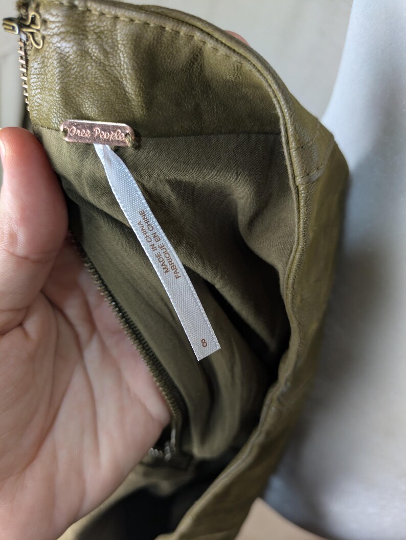 May include: Close-up of an olive green garment with a zipper and a tag that reads "Free People". The tag also includes the text "FABRIQUE EN CHINE". The garment is size 8.