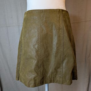 May include: A-line skirt in a muted olive green hue. The skirt is made of a textured material, possibly faux leather, and features vertical seams down the front and back. The skirt is displayed on a mannequin.