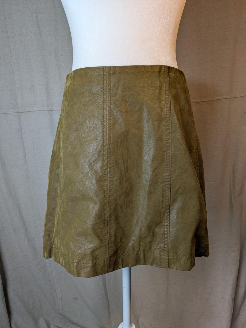 May include: A-line skirt in a muted olive green hue. The skirt is made of a textured material, possibly faux leather, and features vertical seams down the front and back. The skirt is displayed on a mannequin.