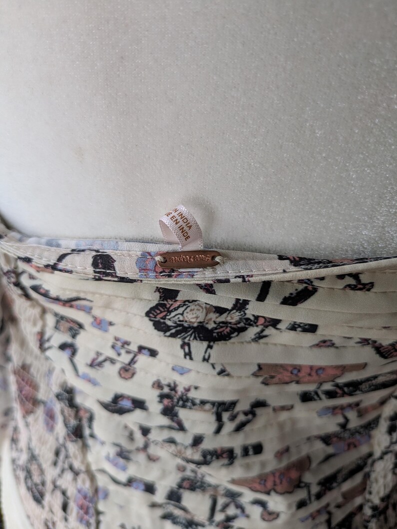 May include: Close-up of a cream-colored garment with a floral pattern in shades of black, pink, and blue. The garment has a tag that reads "MADE IN INDIA" and a small metal tag with the brand name.